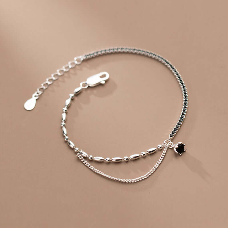 Silver anklet with a small black heart charm and black stones in one side on a beige background