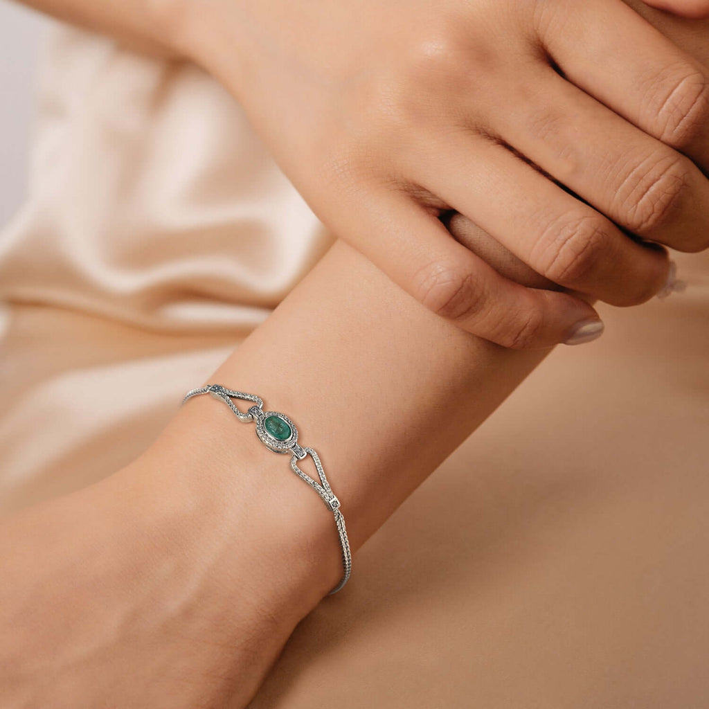 Silver bracelet with a main green emerald gemstone and white zirconia stones on a wrist against a beige background