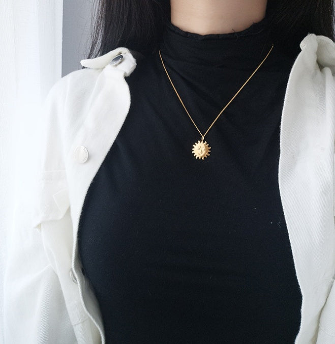Person wearing a black top and white jacket with a gold necklace featuring a sun pendant.