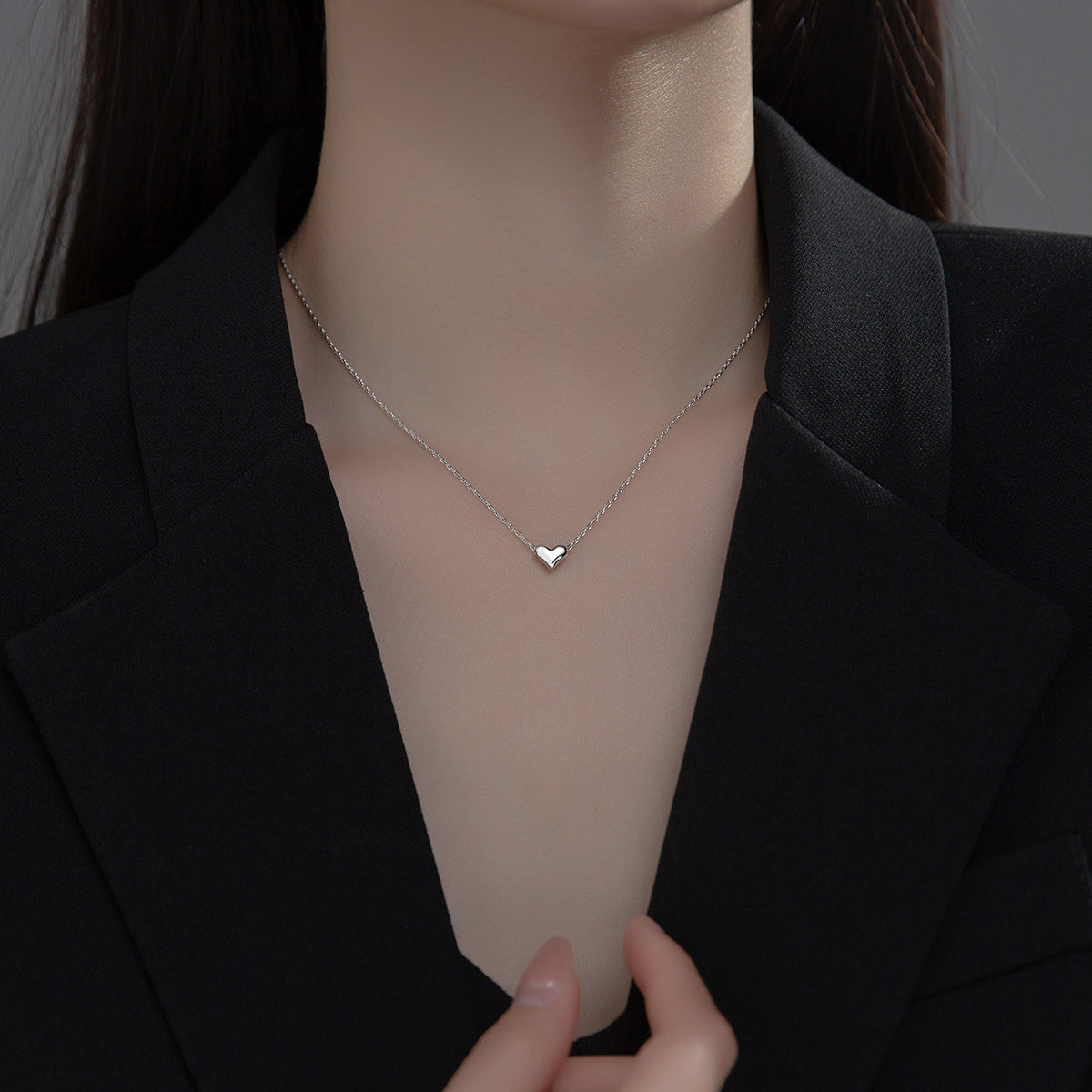Person wearing a delicate silver necklace with a heart pendant against a dark background